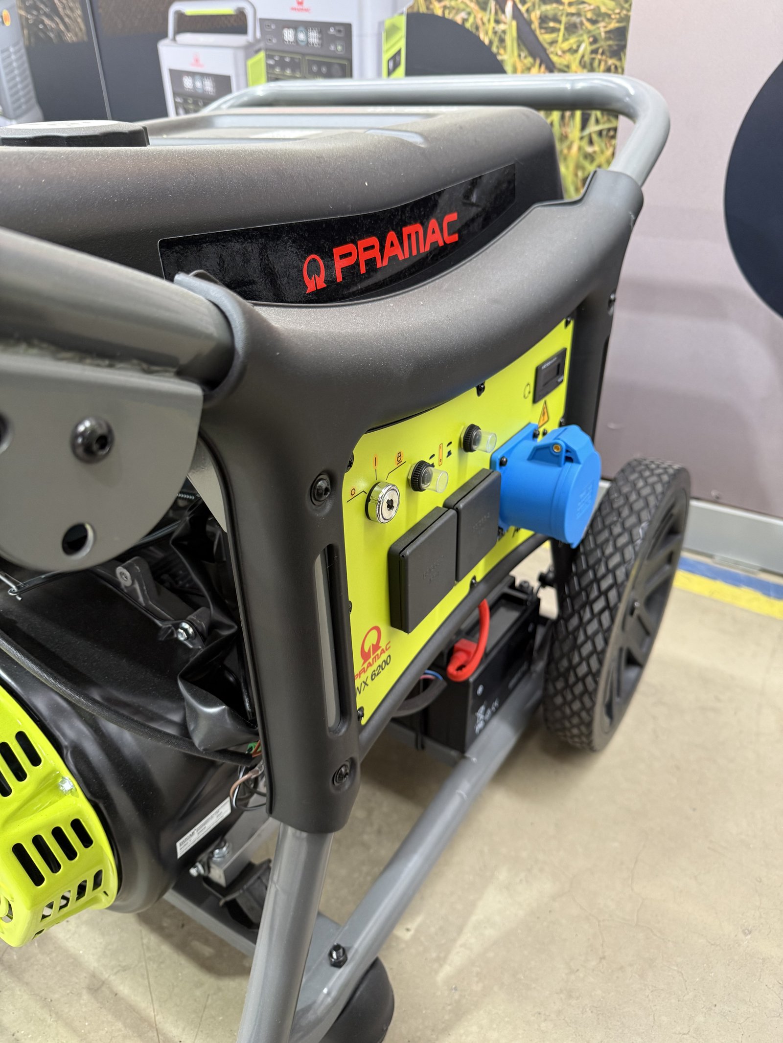 Close-up of a PRAMAC WX 6200 portable petrol generator featuring a heavy-duty tubular steel frame, control panel with 230V blue industrial socket, electric start, and transport wheels. The ge