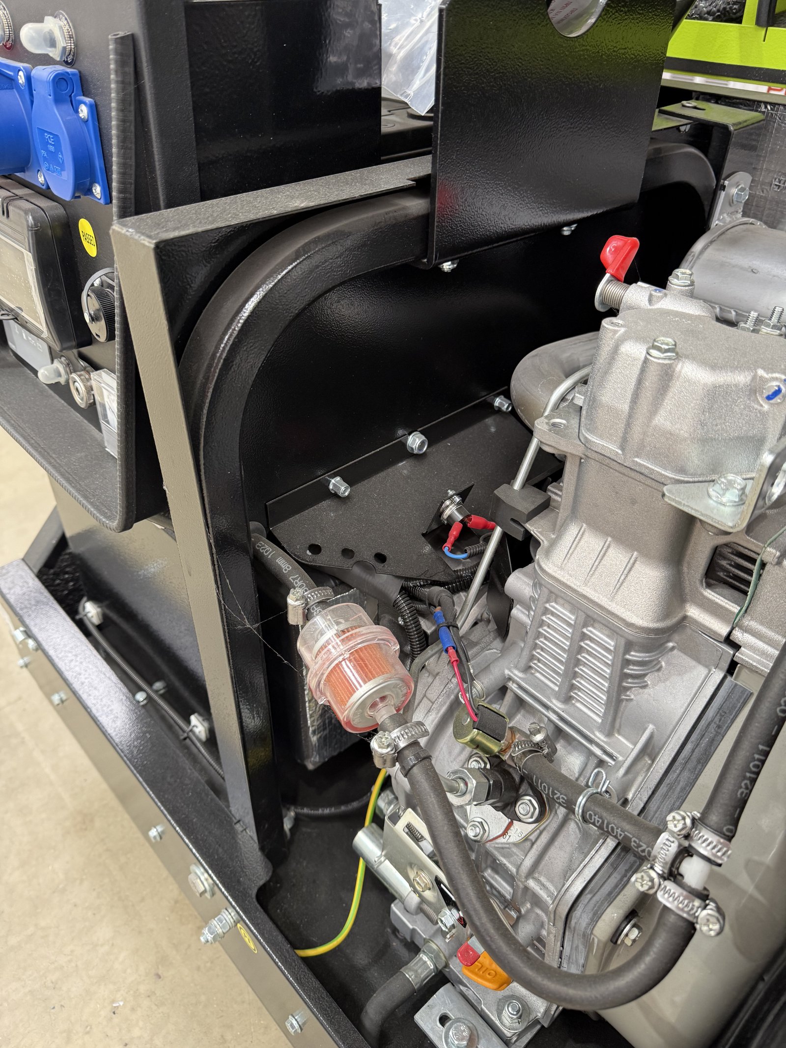 Close-up of the engine compartment on an open-frame petrol generator, showing the engine, fuel filter, fuel lines, wiring, and heavy-duty steel frame. Part of the control panel with industria