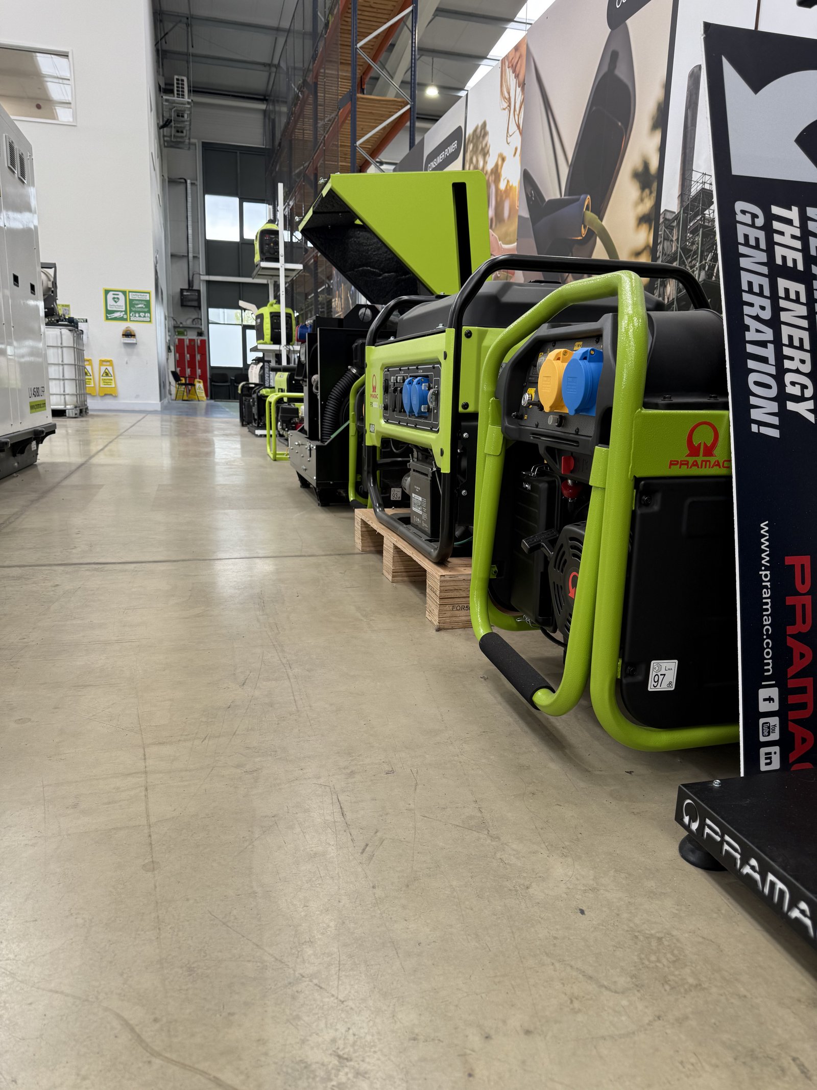 Wide-angle view of multiple PRAMAC portable generators displayed in a modern warehouse showroom. Several open-frame and long-run petrol generators are lined up on pallets, showcasing industri