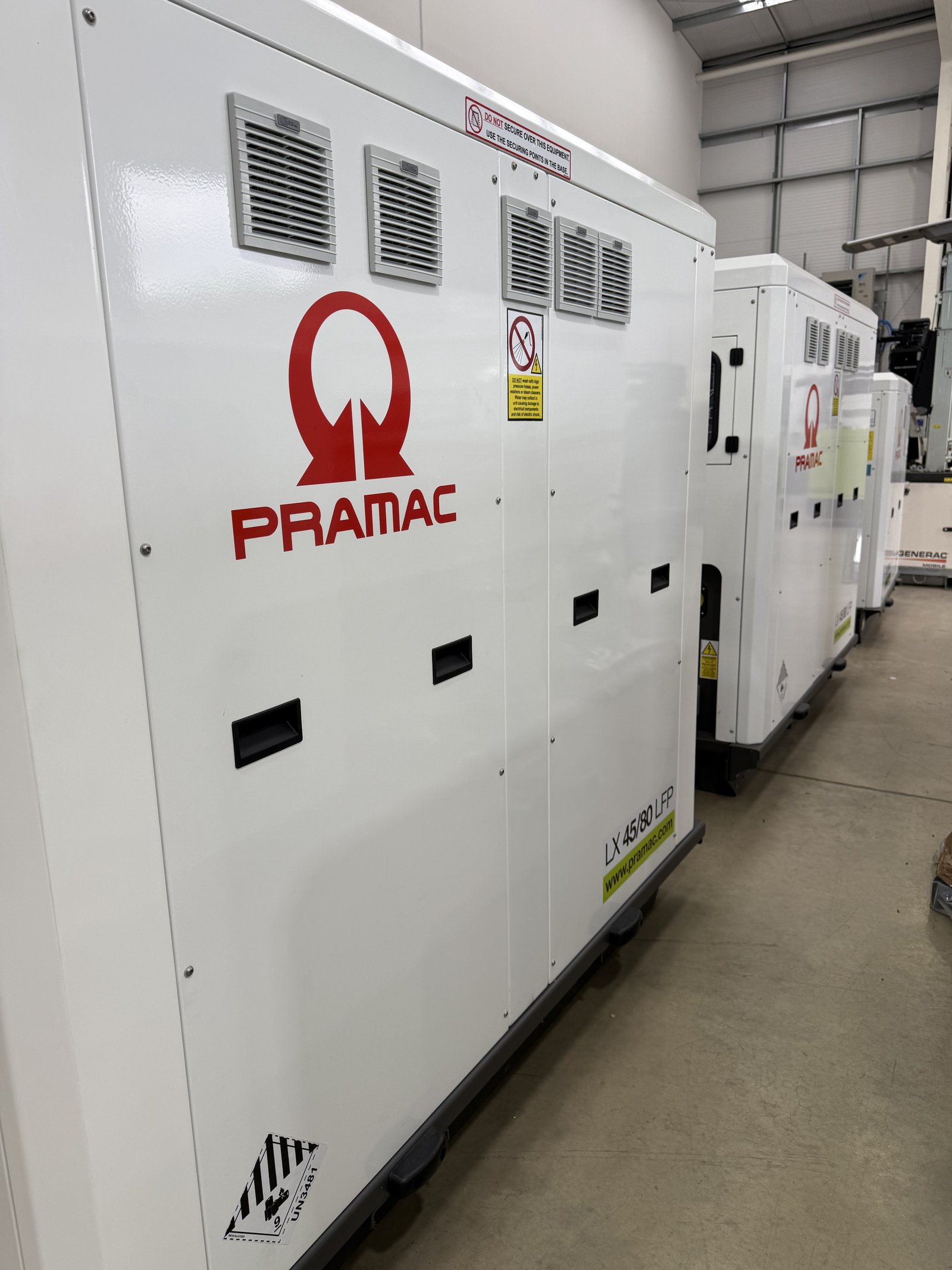 Row of PRAMAC silent diesel generators in white weatherproof acoustic canopies displayed inside a warehouse. The industrial standby generators feature lockable access doors, ventilation grill
