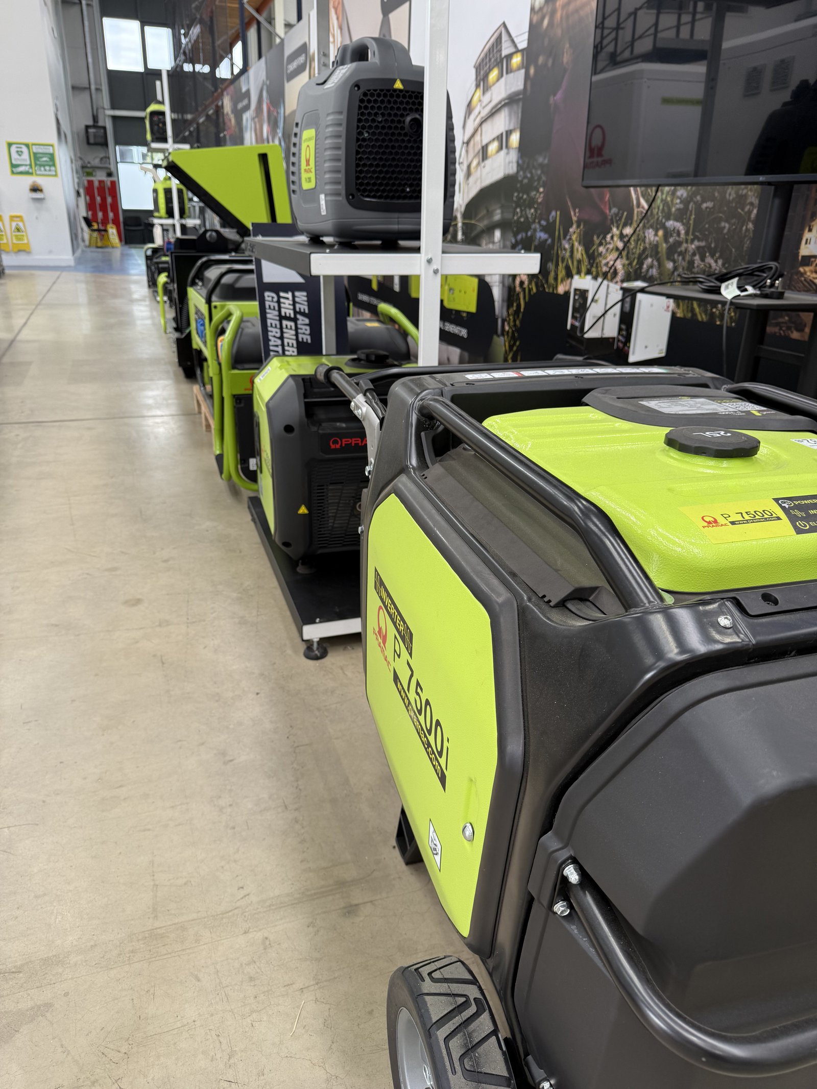 Display of PRAMAC portable inverter and open-frame generators inside a modern warehouse showroom. A PRAMAC P7500i inverter generator is featured in the foreground, with additional portable ge