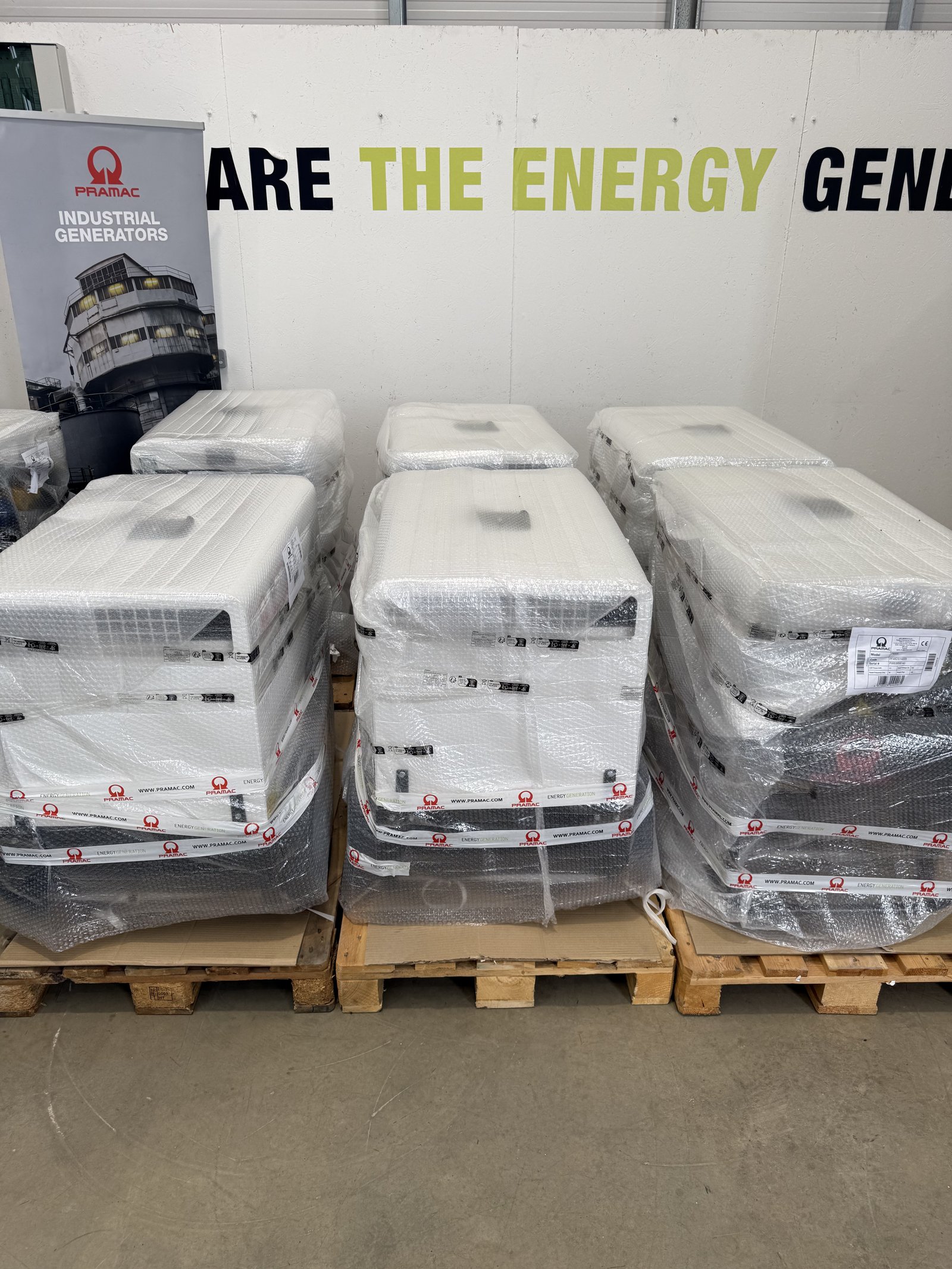 Multiple PRAMAC silent diesel generators securely wrapped and palletised inside a warehouse, ready for dispatch. The weatherproof acoustic generators are packaged for transport, showcasing pr