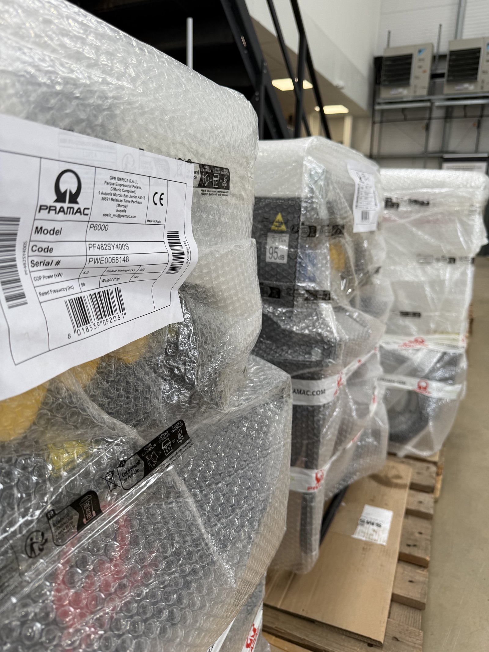 Close-up of multiple PRAMAC P6000 portable generators securely wrapped in protective bubble wrap and palletised for shipment inside a warehouse. Product labels are visible on the packaging, h