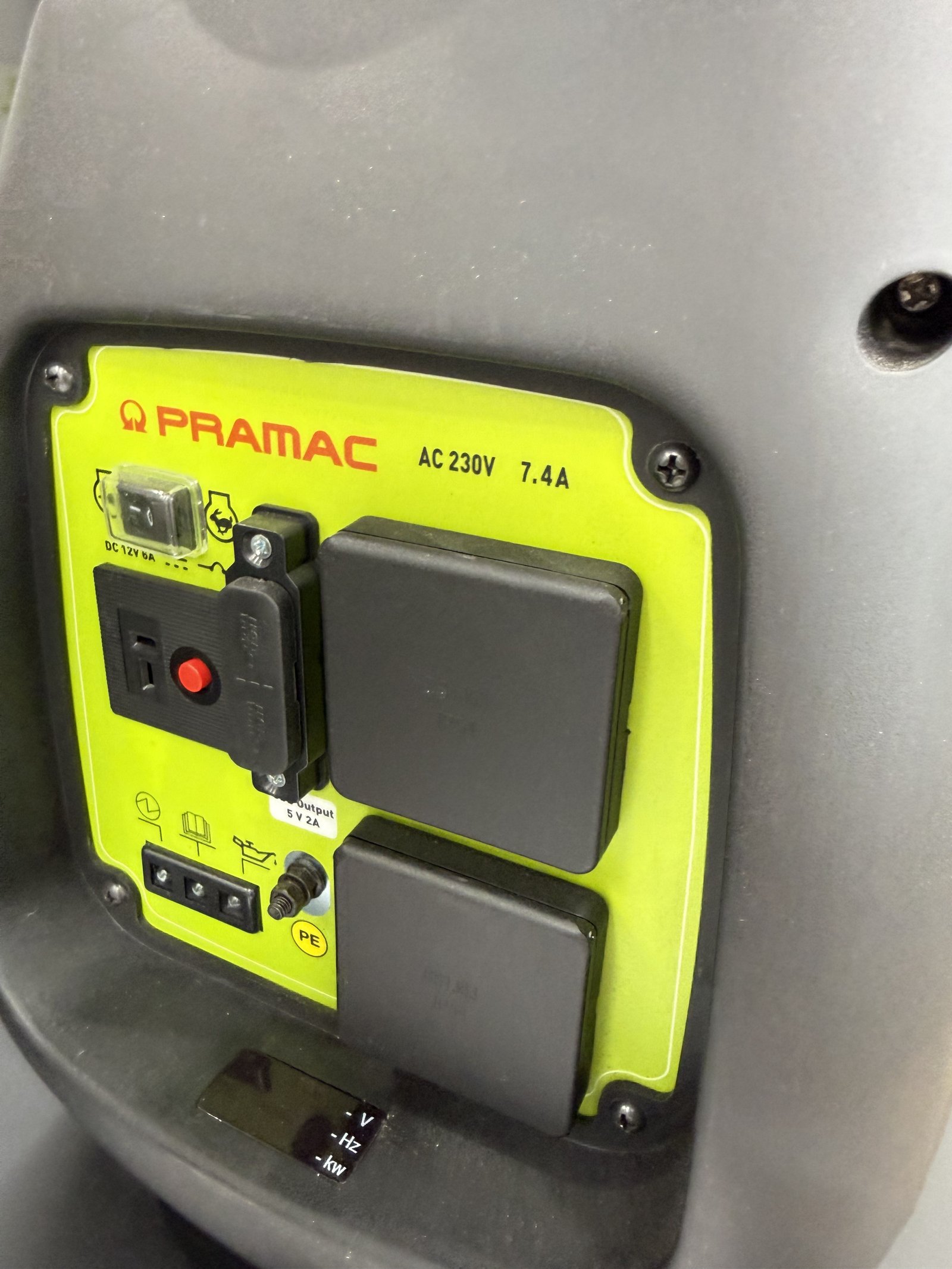 Close-up of the control panel on a PRAMAC portable inverter generator, featuring two covered 230V AC sockets, a 12V DC charging output, USB charging port, circuit breaker, grounding point, an